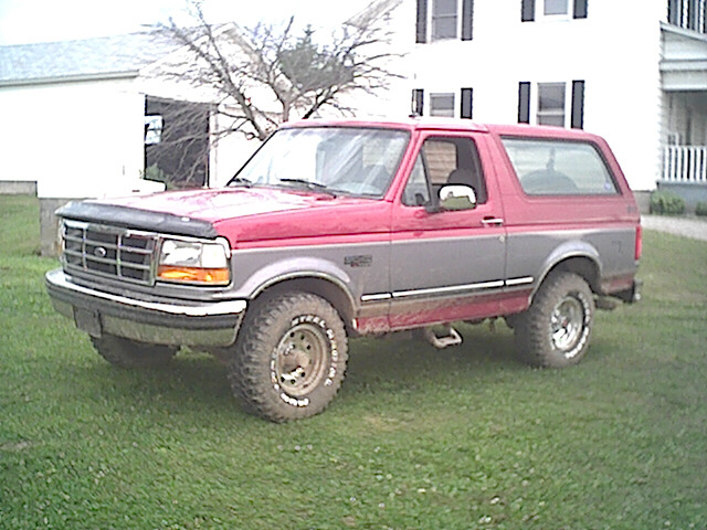 Red and gray Ford Bronco parked on grass in front of a white farmhouse