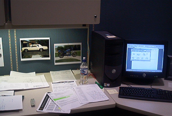Office desk with a computer, keyboard, monitor displaying a spreadsheet, and two framed photos on the wall: one of a white car and one of a blue motorcycle