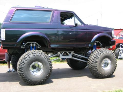 1994 Ford Bronco - This custom lifted Ford Bronco features a unique paint job and oversized tires, with a red truck in the background.