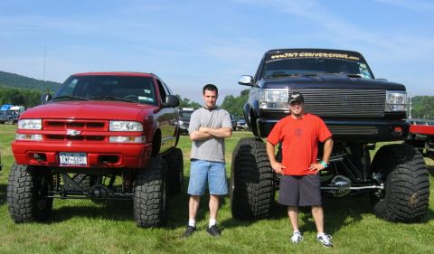 1994 Ford Bronco - Two men pose with their heavily modified monster trucks, a red Chevrolet S-10 and a black Ford Bronco, on a sunny day.