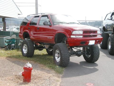 1994 Ford Bronco - This lifted red SUV, likely from the early 2000s, sits high on massive tires, with another lifted vehicle visible to its right.