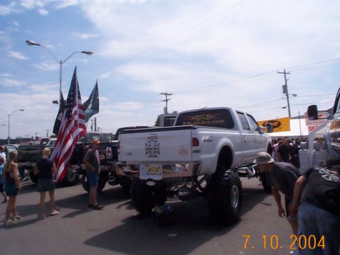 1978 Ford Bronco - A lifted white pickup truck with large tires and American flags is on display at an outdoor event.