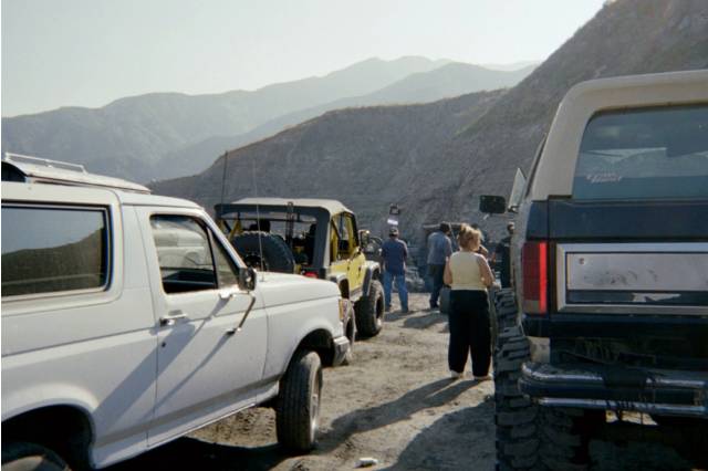 Lineup of full-size SUVs and a yellow Jeep parked off-road with people, mountains behind