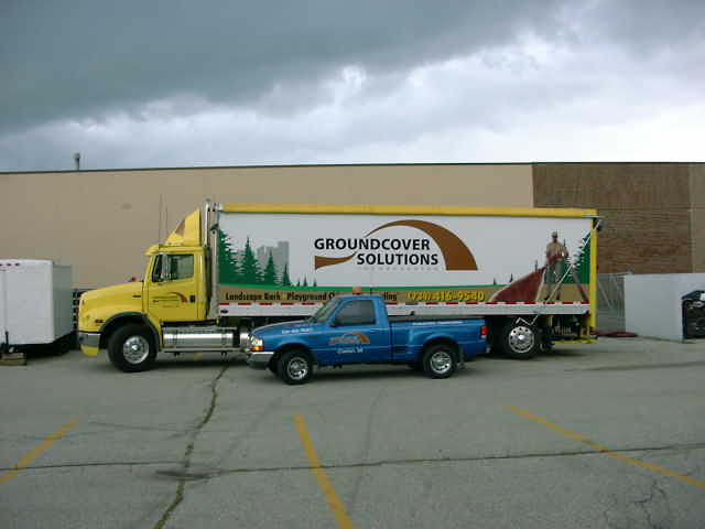 Yellow semi-truck parked next to a blue pickup truck. Semi has 'GROUNDCOVER SOLUTIONS' on its trailer