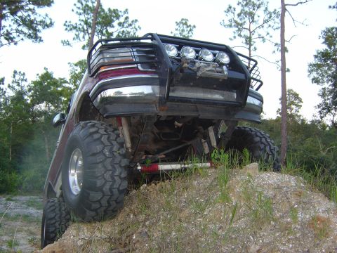 1993 Ford Bronco - Low-angle three-quarter front shot of a lifted maroon Ford 4x4 perched on a sandy mound, shot from the ground so the exposed solid front ax…