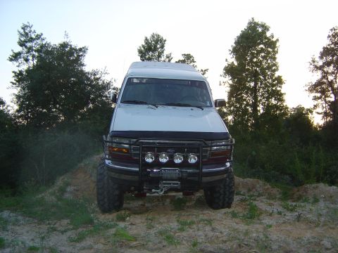 1993 Ford Bronco - Head-on front view of a lifted white-over-burgundy full-size Bronco parked on a sandy rise at dusk, backlit by trees. It wears a black tube…