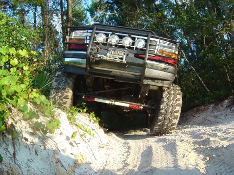 1993 Ford Bronco - Low front-angle view of a lifted 1990s Ford 4x4 cresting a white sand bank, shot from below so the solid front axle, red-boot shocks, steer…