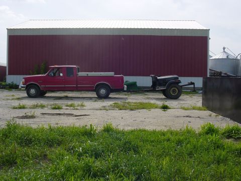 1992 Ford Bronco - A red pickup truck tows a trailer with a black vehicle behind a large red barn.