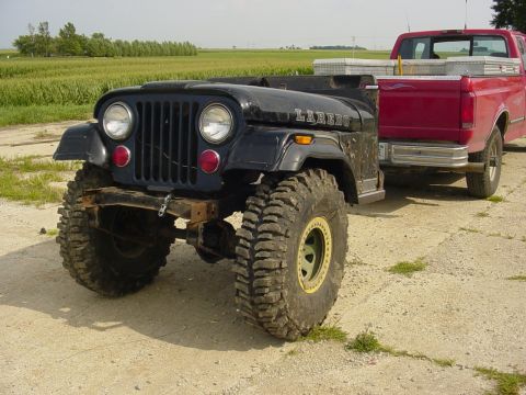 1992 Ford Bronco - A black Jeep CJ-7 Laredo with large off-road tires is being towed by a red pickup truck in a rural setting.