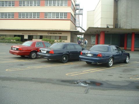 1999 Ford Crown Victoria - Three Ford Crown Victorias are parked outside a medical building, with two men standing near the entrance.