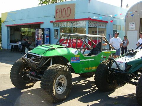 1990 Ford Bronco - Two custom off-road vehicles are parked in front of a building with a 'FOOD' sign and festive bunting.