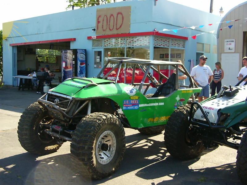 Green off-road buggy with roll cage parked outside a building with a 'FOOD' sign. Another buggy is partially visible to the right