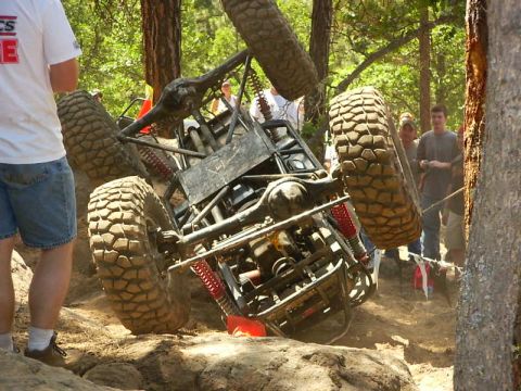 1990 Ford Bronco - An off-road buggy is seen tipped on its side during an event in a wooded area with spectators watching.
