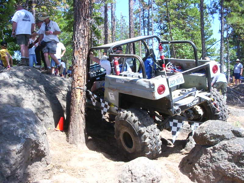 Off-road buggy with checkerboard flag attached to rear bumper, navigating a rocky course. License plate reads 323-KPU