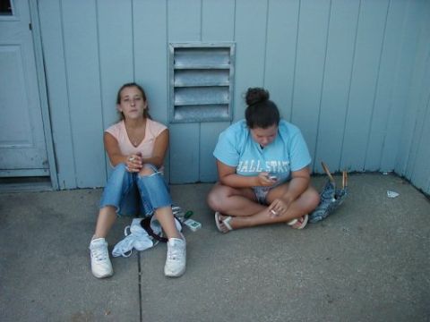 1997 Ford Taurus - Two young women sit on the ground next to a blue building, one looking at her phone and smoking.