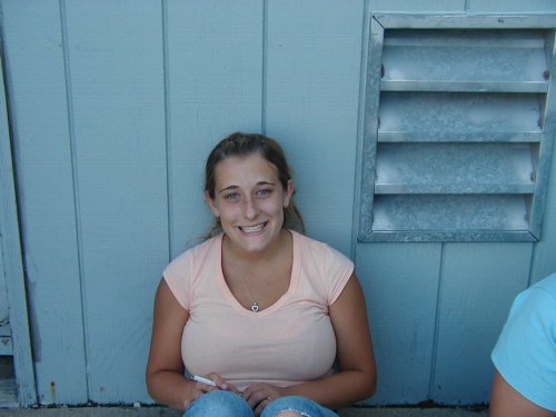 Woman in a peach t-shirt and jeans sits in front of a blue wall with a metal vent. Supermotors.net logo visible