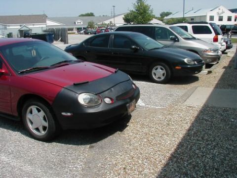 1997 Ford Taurus - A red car with a car bra is parked next to other vehicles in a gravel lot.