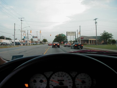 Dashboard view from driver's seat looking at a road with multiple cars, a Sears Auto Center sign, and utility poles