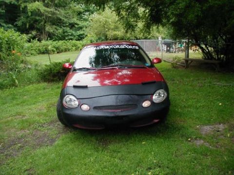 1997 Ford Taurus - A red 1997 Ford Taurus is parked in a grassy yard, partially covered by a black car bra and a 'NORTON MOTORS SPORTS' banner on the windshie…