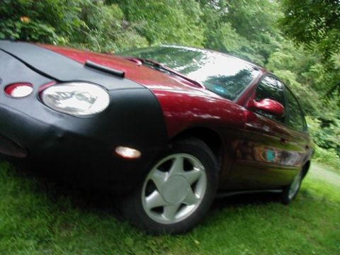 1997 Ford Taurus - A red 1997 Ford Taurus is partially covered by a black car bra, parked outdoors on a grassy area.
