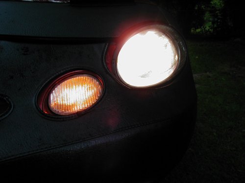Car headlight and amber turn signal illuminated, covered by a dark car bra