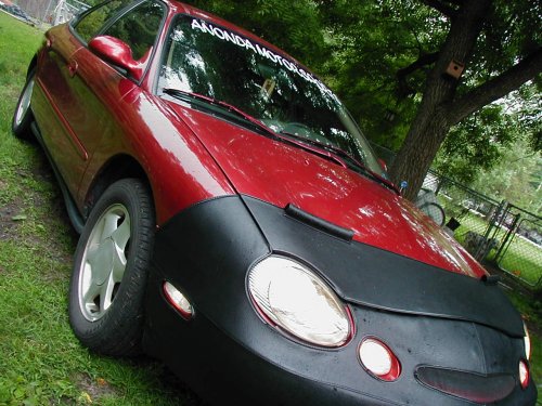 red sedan with a car bra, parked on grass near a tree. windshield banner reads ANONDA MOTORSPORTS. Supermotors.net logo on lower bumper