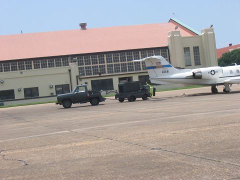 1988 Ford Bronco - A green pickup truck tows a trailer across an airport tarmac, with a jet and building in the background.