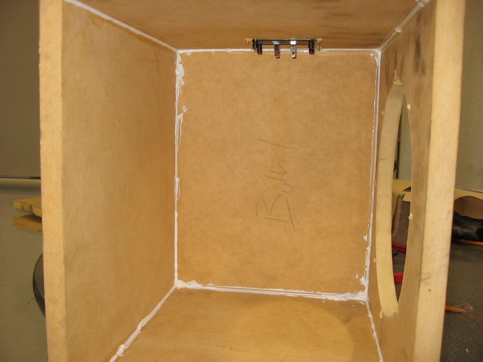 Interior of an unfinished wooden speaker box with sealant visible on the edges. Some writing is scrawled on the back panel