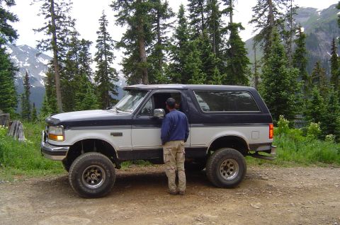 1993 Ford Bronco - A lifted, two-tone Ford Bronco is parked on a dirt road with a mountainous, forested background.