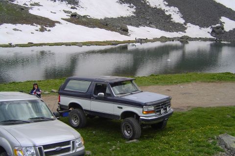 1993 Ford Bronco - A lifted Ford Bronco sits by a lake with a snowy mountain backdrop.