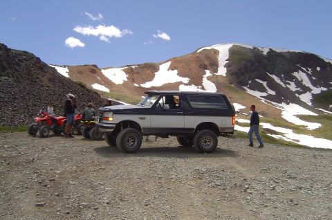 1993 Ford Bronco - A two-tone Ford Bronco is parked on a dirt road, with ATVs nearby and mountains in the distance.