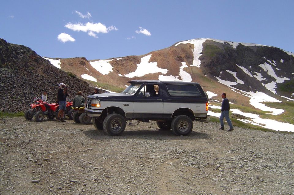 Blue and white lifted Ford Bronco on a gravel mountain pass with snowy peak behind