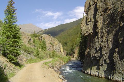 1993 Ford Bronco - Mountain landscape with a dirt road, river, and pine trees under a blue sky.