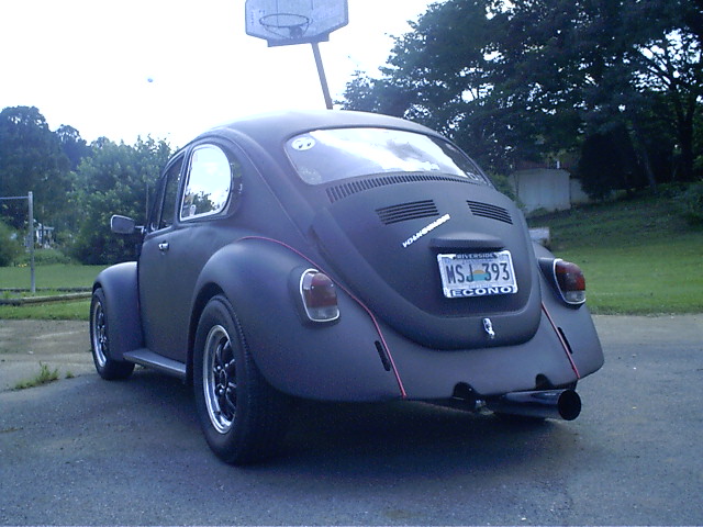Rear three-quarter view of a flat-black VW Beetle with chrome wheels and a side-exit exhaust on a driveway