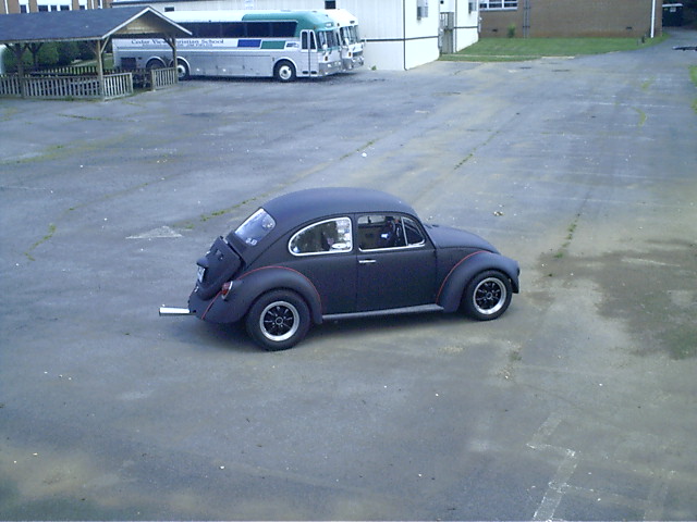 Flat-black VW Beetle sitting in a large empty lot with vintage buses parked along the back fence