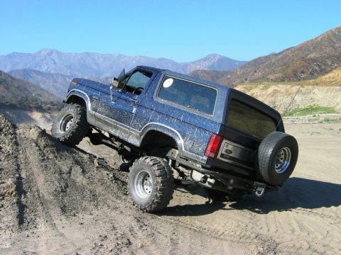 1985 Ford Bronco - A blue Ford Bronco navigates a steep dirt incline in a mountainous, arid landscape.