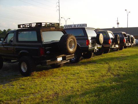 1986 Ford Bronco - A row of black Ford Broncos, including a 1986 model with a roof rack and spare tire, are parked on a grassy lot.