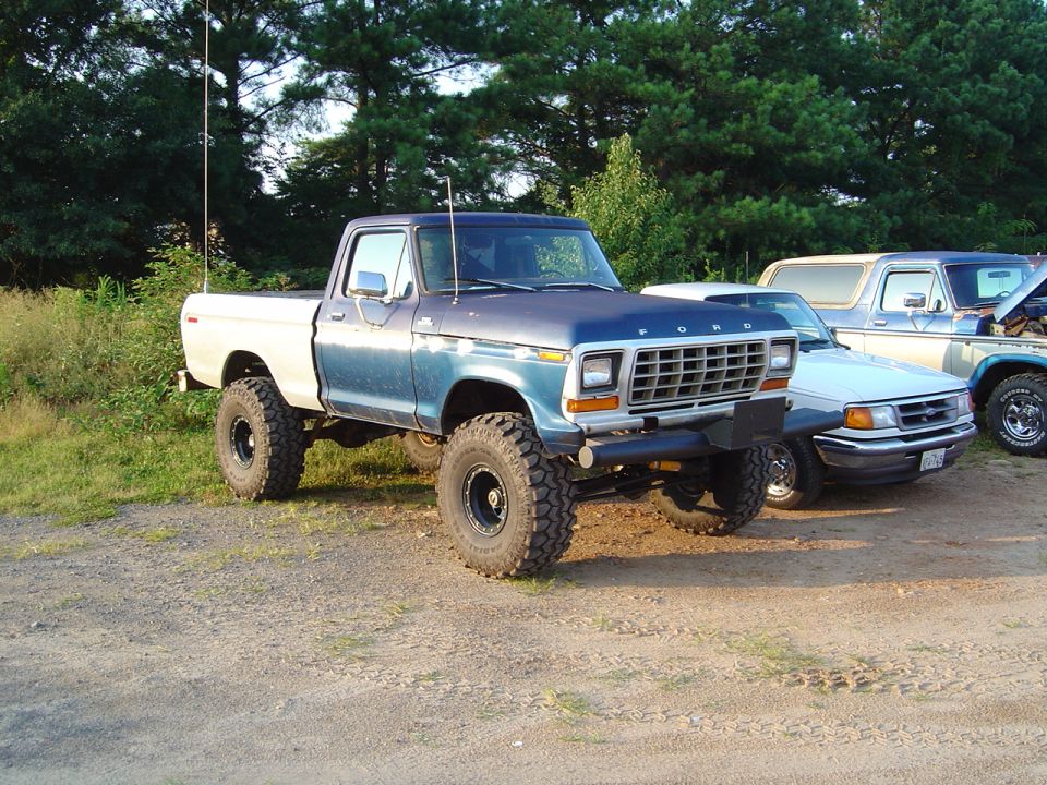Lifted 1979 Ford F-Series pickup truck with aftermarket wheels and tires. Another Ford Bronco and pickup truck are parked behind it