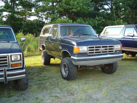 1986 Ford Bronco - A dark blue Ford Bronco with a lifted suspension is parked on grass next to another vehicle.