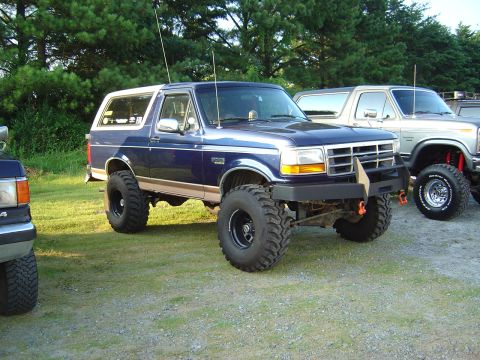 1986 Ford Bronco - This lifted Ford Bronco is parked next to another similar vehicle outdoors.