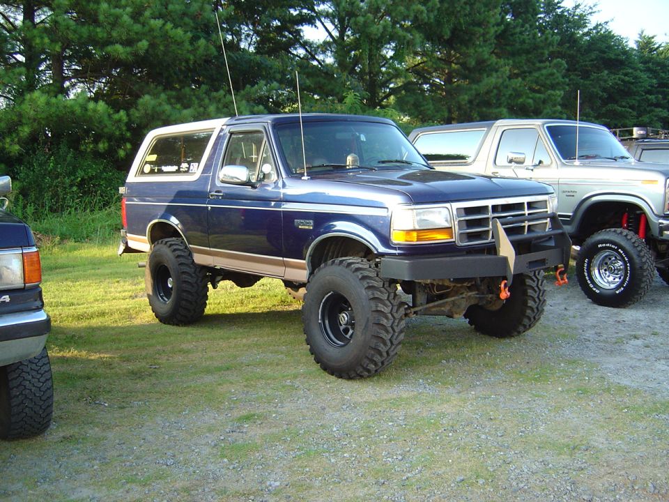 Blue lifted Ford Bronco with aftermarket bumper and large tires. Another Bronco is parked next to it