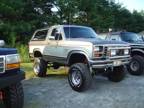 1986 Ford Bronco - A lifted Ford Bronco is parked next to other trucks outdoors.