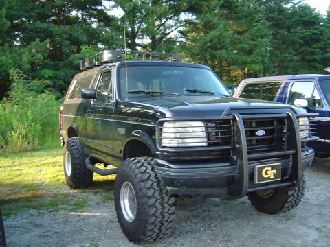 1986 Ford Bronco - Black lifted OBS-body Ford Bronco parked on gravel at the edge of a treeline, fitted with a full black brush guard, roof rack with light ba…