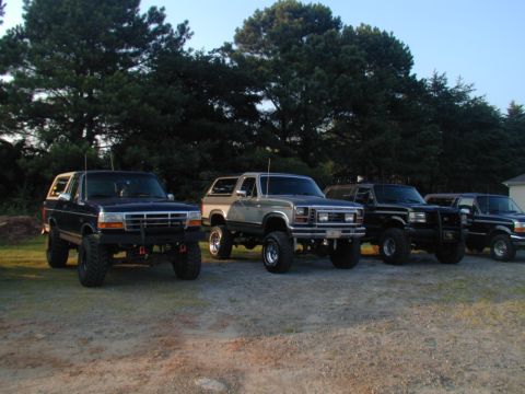 1988 Ford Bronco - A lineup of four lifted Ford Broncos shows off their off-road modifications under a clear sky.