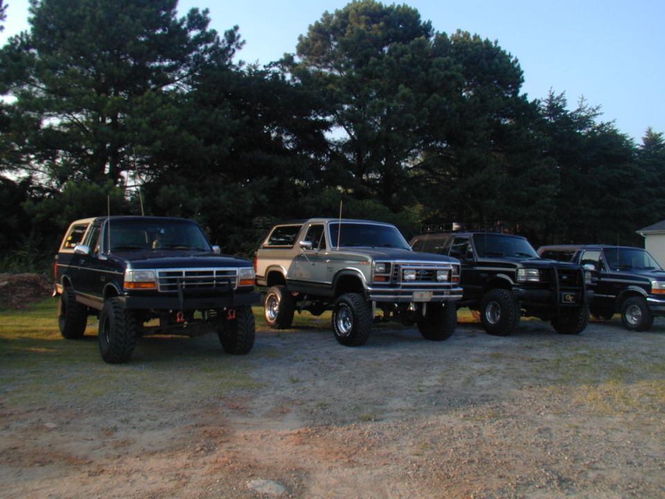 Four lifted Ford Broncos parked on gravel, trees in background. The first two are dark blue and silver/gray. The last two are black