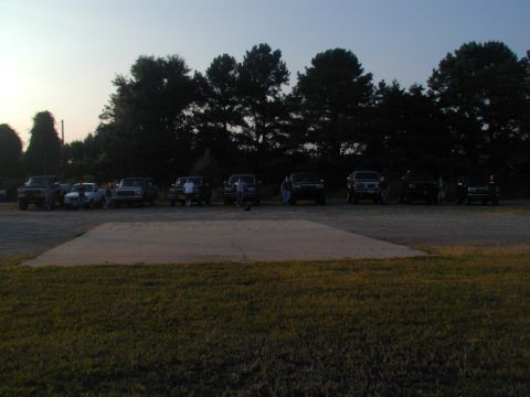 1988 Ford Bronco - A group of SUVs and trucks are parked in a lot as the sun sets, with a concrete pad in the foreground.