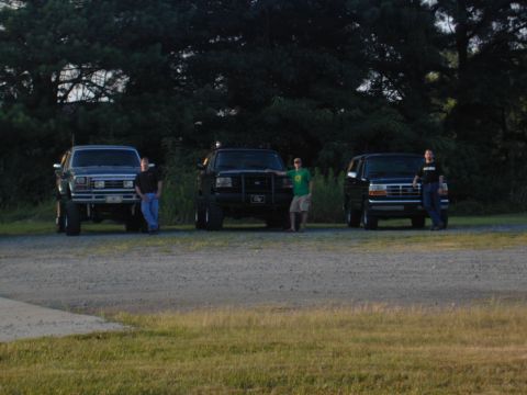1988 Ford Bronco - Three trucks and an SUV are parked on a gravel lot, with individuals standing next to each vehicle.