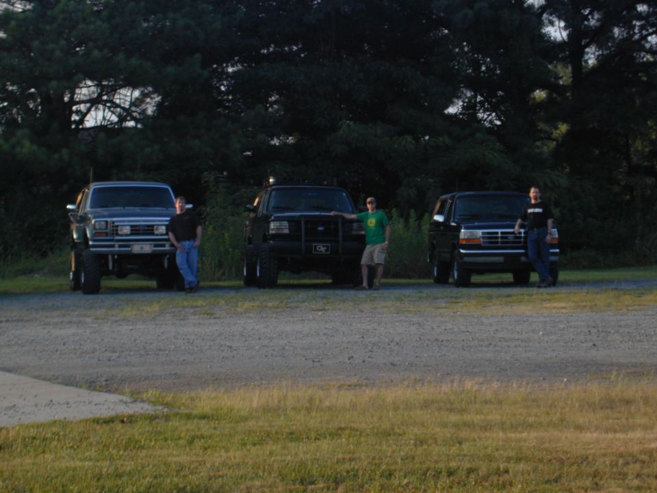 Three trucks and an SUV parked on a gravel lot; the leftmost truck is a 1987 Ford Bronco