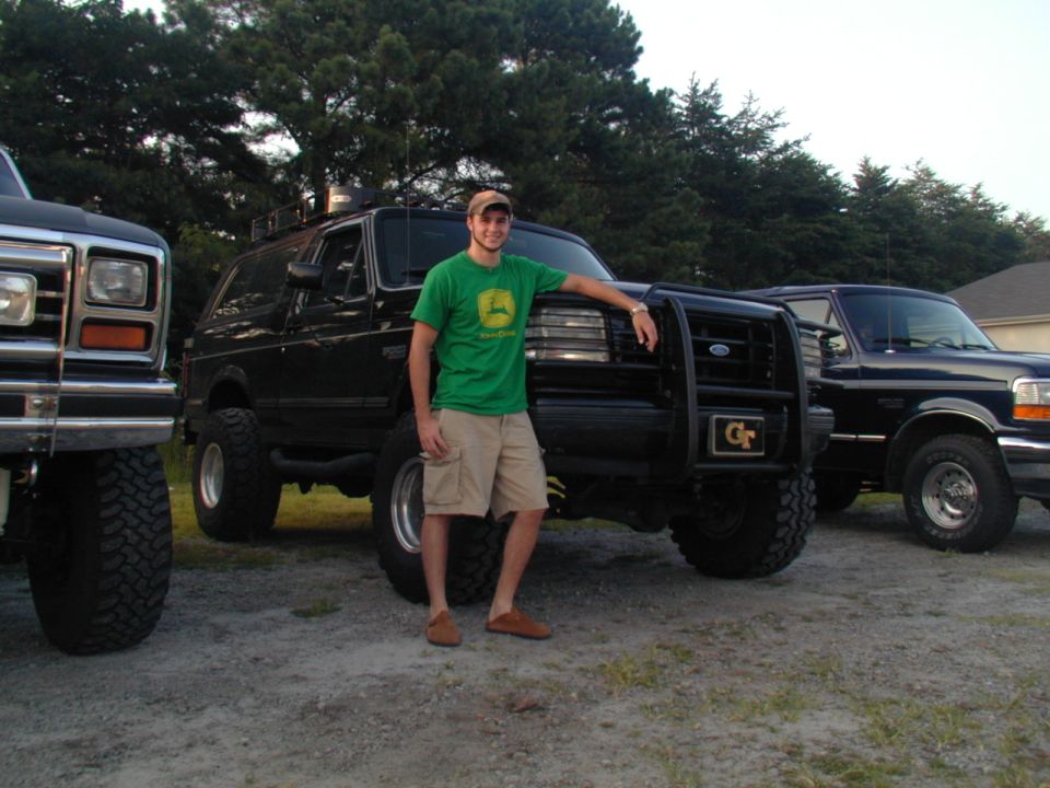 Man standing next to a lifted 1988 Ford Bronco with a brush guard; another Ford truck is to the left, and a third to the right