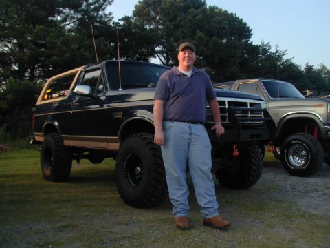 1988 Ford Bronco - A man stands beside a lifted Ford Bronco, with another truck visible behind it.